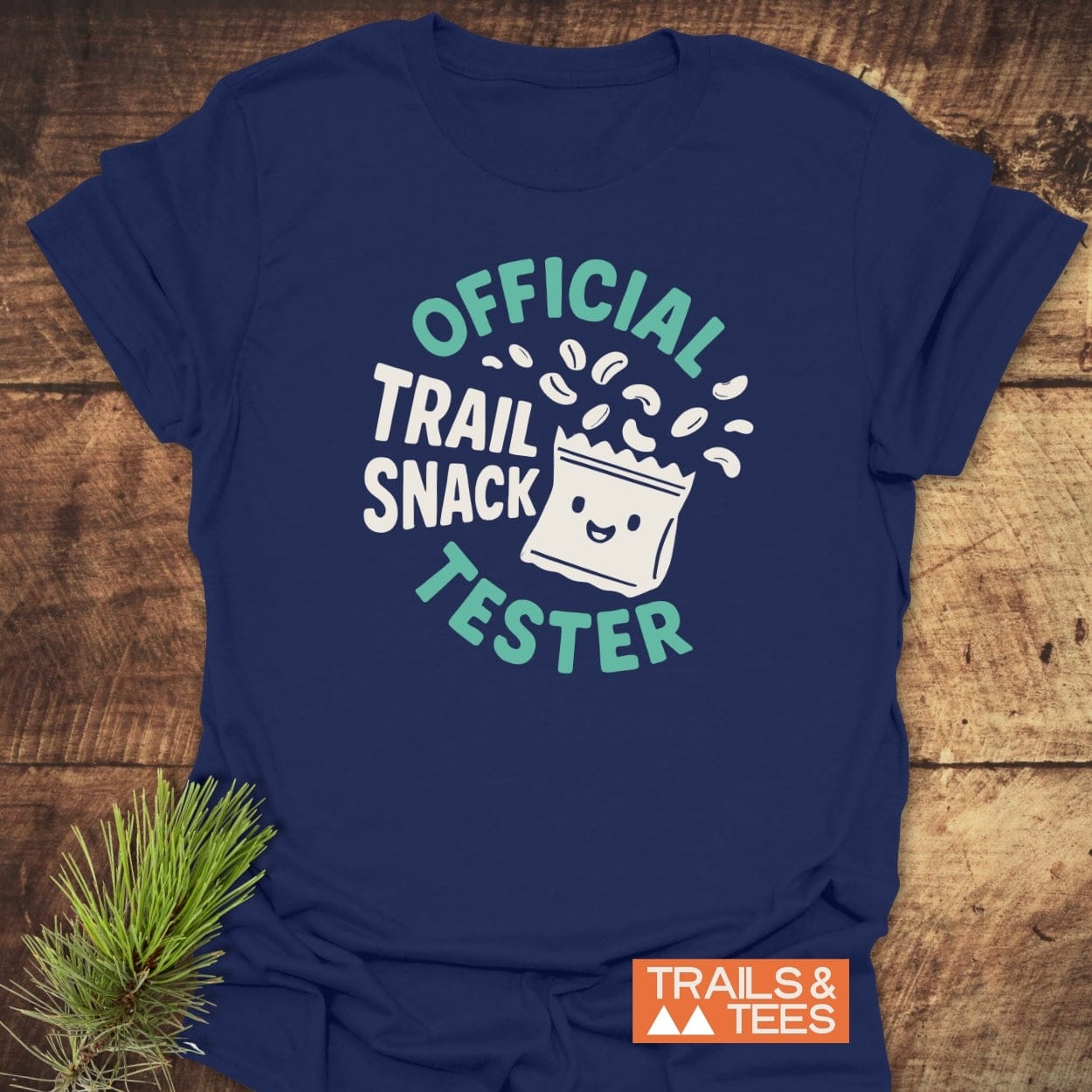 Navy-blue Official Trail Snack Tester T-shirt with a cute trail-mix bag graphic, featuring durable DTF prints and an orange TRAILS & TEES logo, laid on a wooden surface.