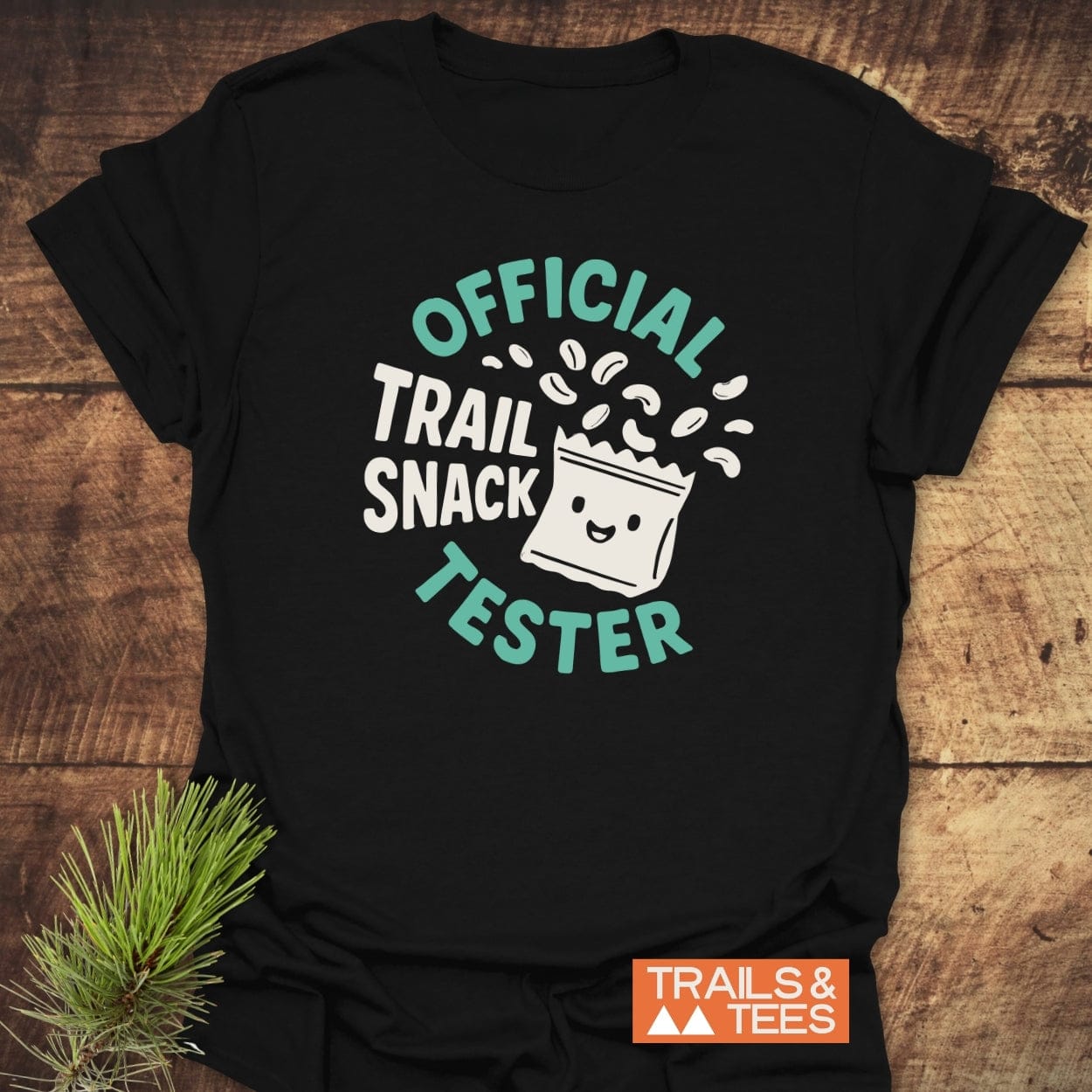 Official Trail Snack Tester T-Shirt featuring a cartoon snack bag and text, laid on wood. Displays TRAILS & TEES logo. Made of soft ring-spun cotton for outdoor enthusiasts.