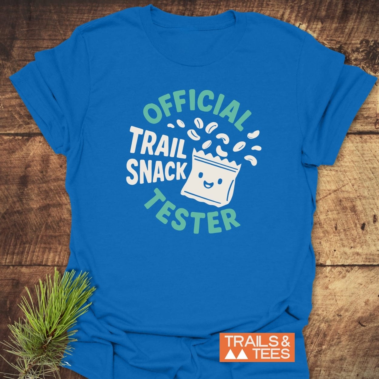 Official Trail Snack Tester T-Shirt featuring a smiling snack-bag design with trail-mix bits, laid on a wooden surface. Includes TRAILS & TEES logo and pine sprig styling prop.