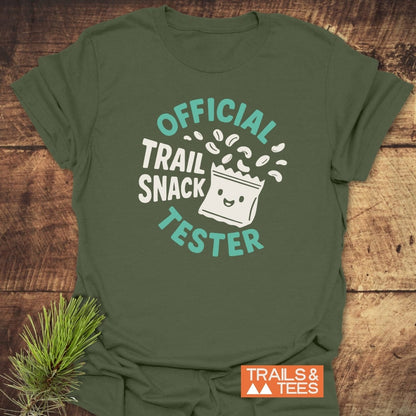 Official Trail Snack Tester T-Shirt featuring a playful trail-mix design, laid on rustic wood with a pine sprig. Includes Trails & Tees logo, highlighting its outdoor-loving spirit and comfortable ring-spun cotton material.
