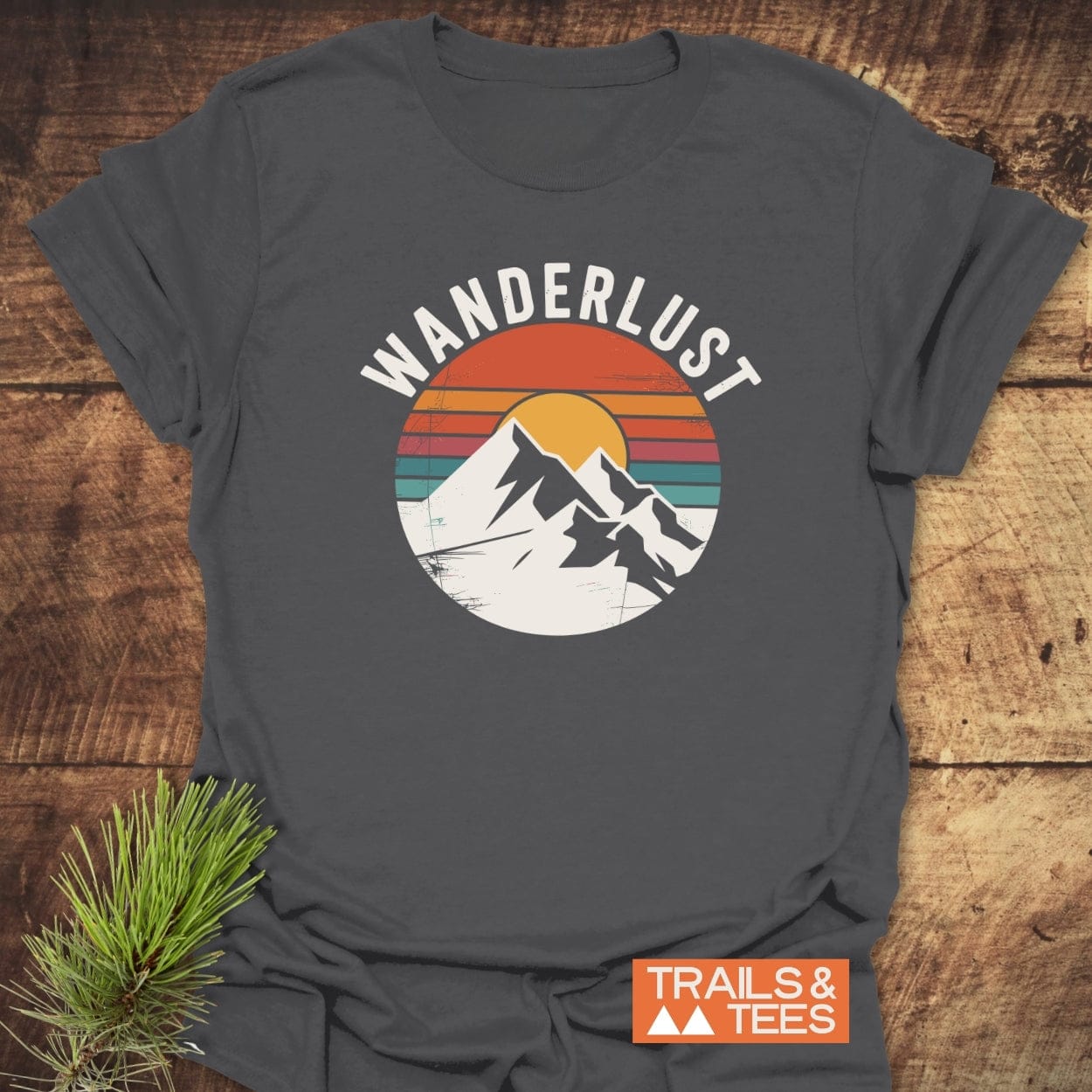 Wanderlust Retro T-Shirt featuring a circular graphic of snow-capped mountains against a striped sunset with WANDERLUST above. Dark gray design, styled with a pine sprig and TRAILS & TEES logo.