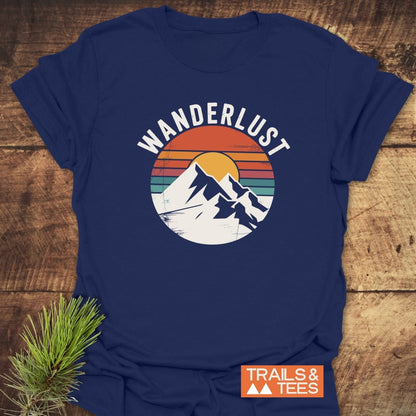 Wanderlust Retro T-Shirt featuring a mountain and sunset graphic, vibrant retro stripes, and “WANDERLUST” text. Includes “TRAILS & TEES” label. Displayed on rustic wood with pine sprig.