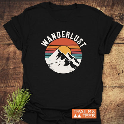 Wanderlust Retro T-Shirt featuring snowy mountain peaks and a setting sun graphic, laid on wood. Includes “TRAILS & TEES” logo, emphasizing adventure-ready, soft ring-spun cotton design.