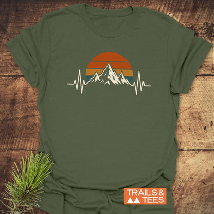 Mountain Heartbeat T-Shirt displayed on rustic wood, featuring a mountain and retro sun graphic with EKG lines. A pine sprig and orange TRAILS & TEES label accentuate the outdoor-inspired design.