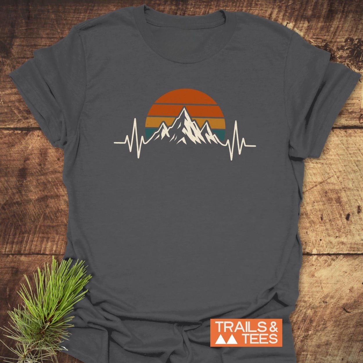 Mountain Heartbeat T-Shirt features a mountain-to-heartbeat graphic with a retro sunset on soft charcoal gray cotton, perfect for outdoor enthusiasts. Includes “TRAILS & TEES” label, ensuring comfort and durability.