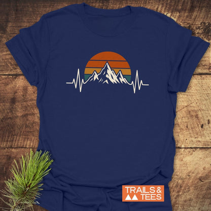 Mountain Heartbeat T-Shirt featuring stylized mountain peaks against a retro sunset with EKG line, laid on wooden surface. Includes TRAILS & TEES logo and pine sprig for outdoorsy vibe.
