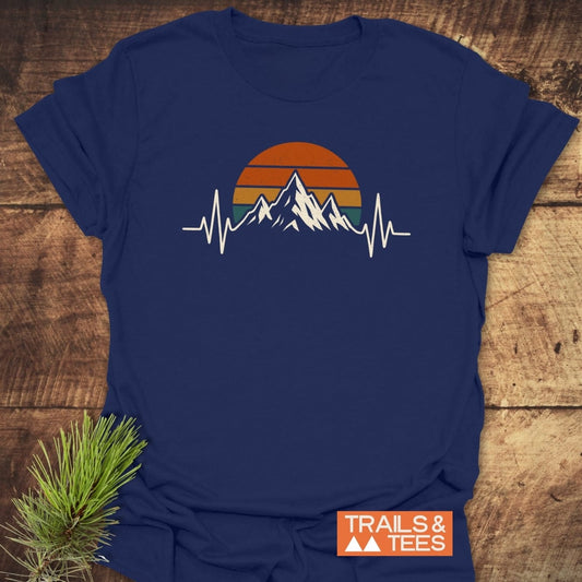 Mountain Heartbeat T-Shirt featuring stylized mountain peaks against a retro sunset with EKG line, laid on wooden surface. Includes TRAILS & TEES logo and pine sprig for outdoorsy vibe.