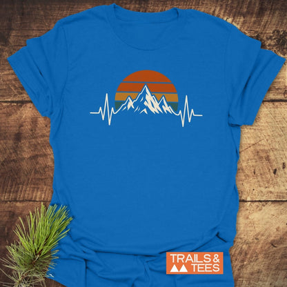 Mountain Heartbeat T-Shirt featuring a mountain silhouette and sunset graphic with a white heartbeat line, laid on wood. A “TRAILS & TEES” label and pine sprig accent the scene.