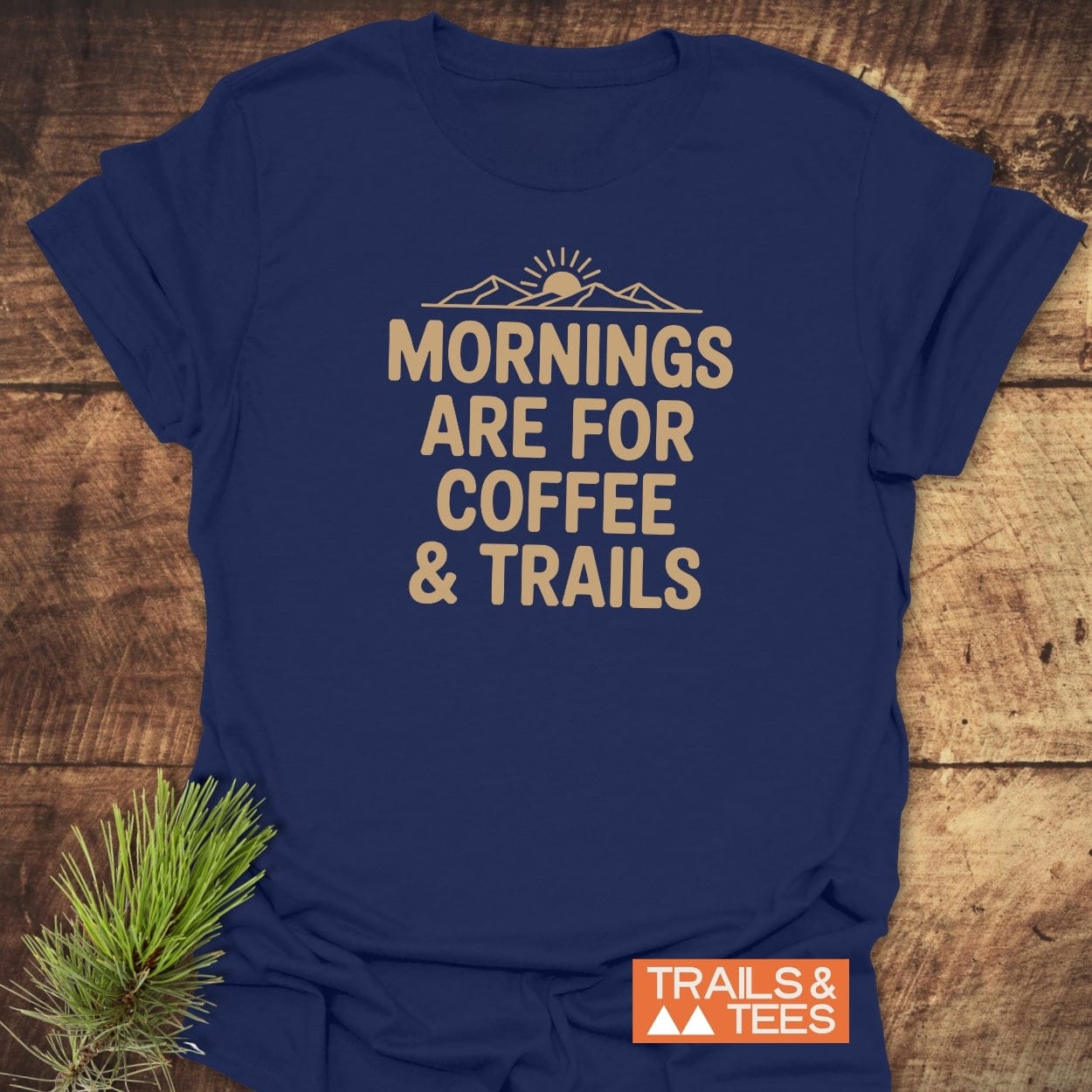 Morning Coffee & Trails T-Shirt features a sun and mountain graphic with MORNINGS ARE FOR COFFEE & TRAILS slogan. Lying on wood, it includes a pine sprig and TRAILS & TEES logo.