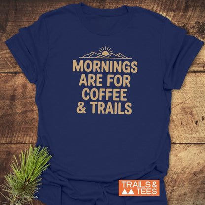Morning Coffee & Trails T-Shirt features a sun and mountain graphic with MORNINGS ARE FOR COFFEE & TRAILS slogan. Lying on wood, it includes a pine sprig and TRAILS & TEES logo.