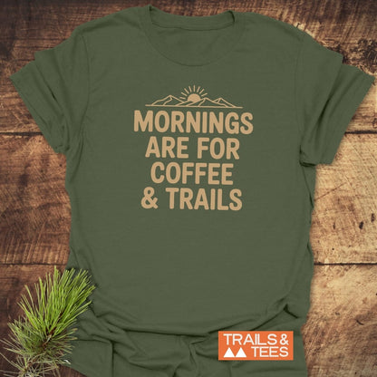 CMorning Coffee & Trails T-Shirt featuring a rising sun and mountain graphic with the slogan. Laid on wood, a pine sprig adds rustic charm, showcasing its adventure-ready design.