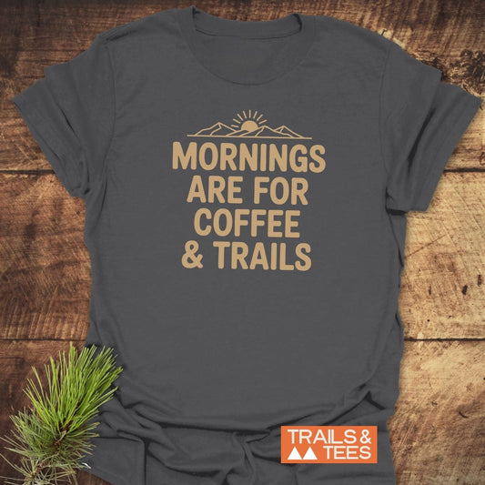 CMorning Coffee & Trails T-Shirt featuring a line-art sunrise and slogan, laid on wood, with a rustic pine sprig, highlighting outdoor adventure and comfort from durable, pre-shrunk, ring-spun cotton.