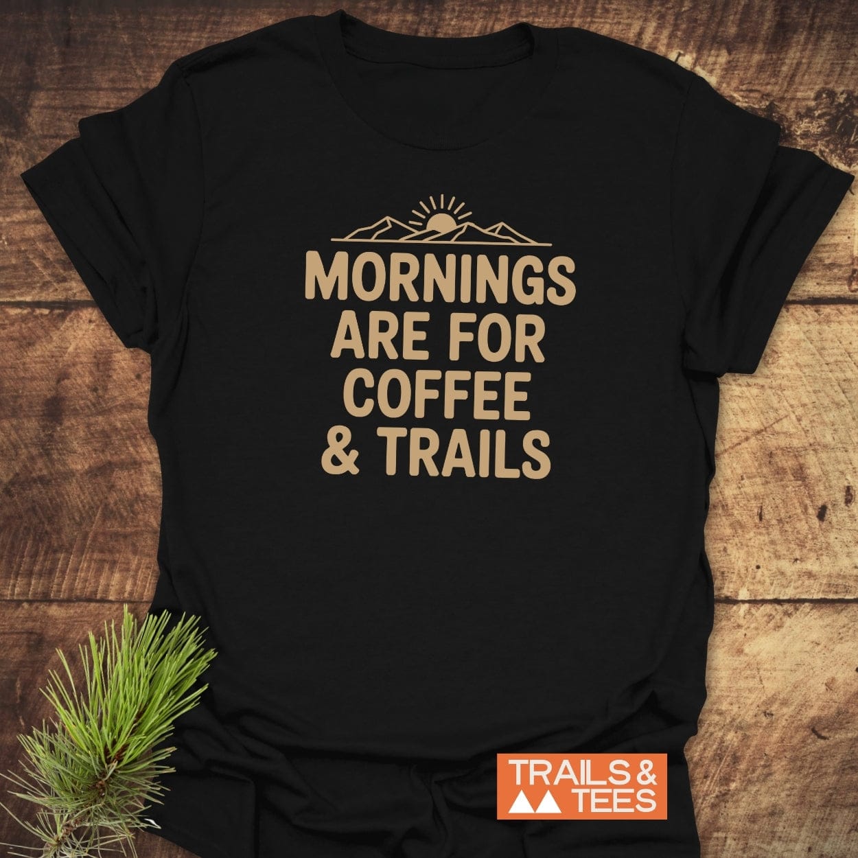 CMorning Coffee & Trails T-Shirt featuring beige text and a mountain graphic on black fabric, laid flat with a pine branch beside it; embodies outdoor spirit and adventure-ready comfort.