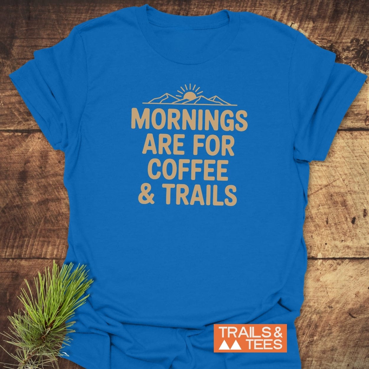 CMorning Coffee & Trails T-Shirt flat-lay shows tan lettering and sunrise graphic on blue fabric, laid on wood with pine sprig and 'TRAILS & TEES' logo tag, reflecting outdoor adventure spirit.