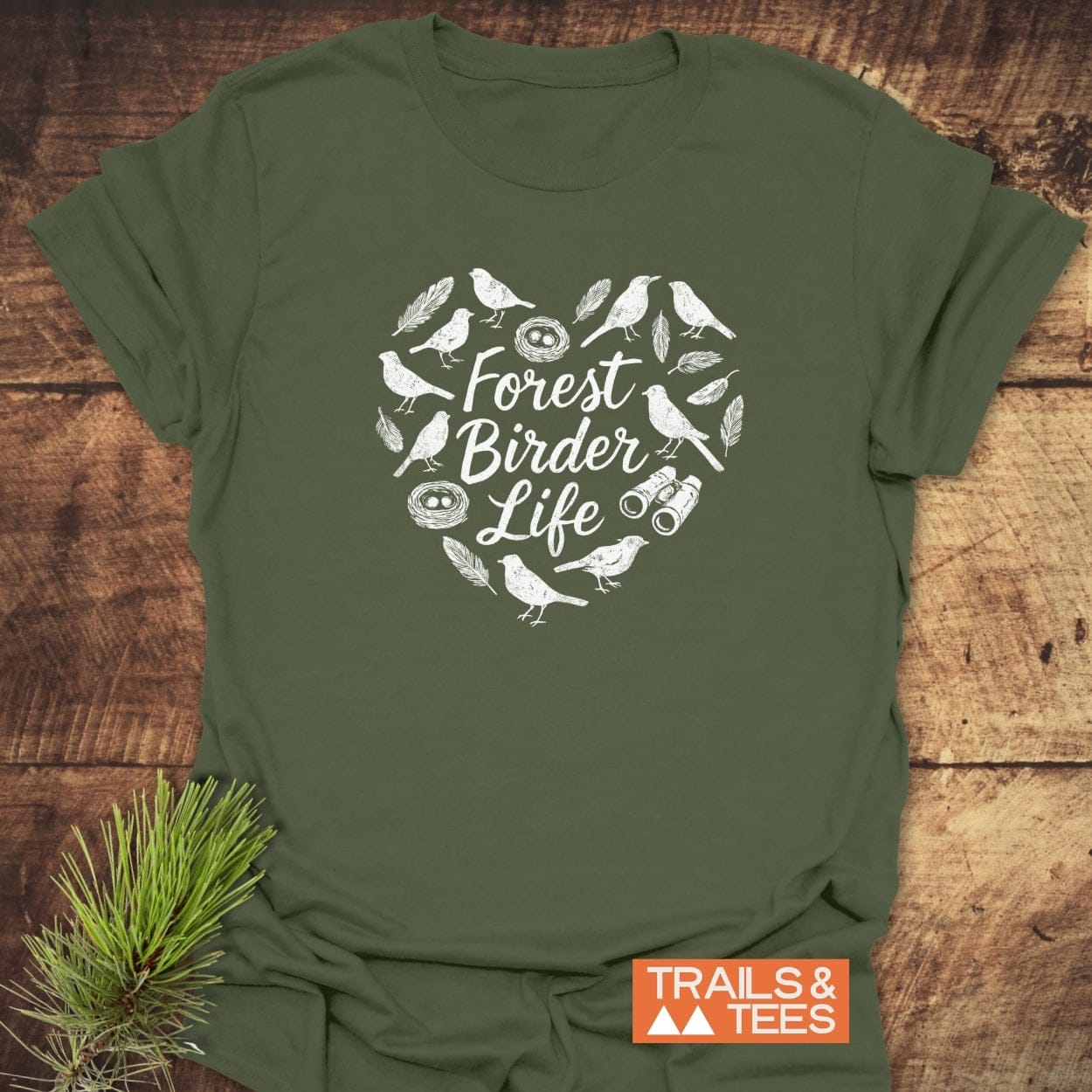Forest Birder Life T-Shirt with a heart-shaped nature graphic, featuring birds, feathers, binoculars, and nests. The soft, olive-green tee lies flat, accented by pine needles, embodying outdoor adventure.