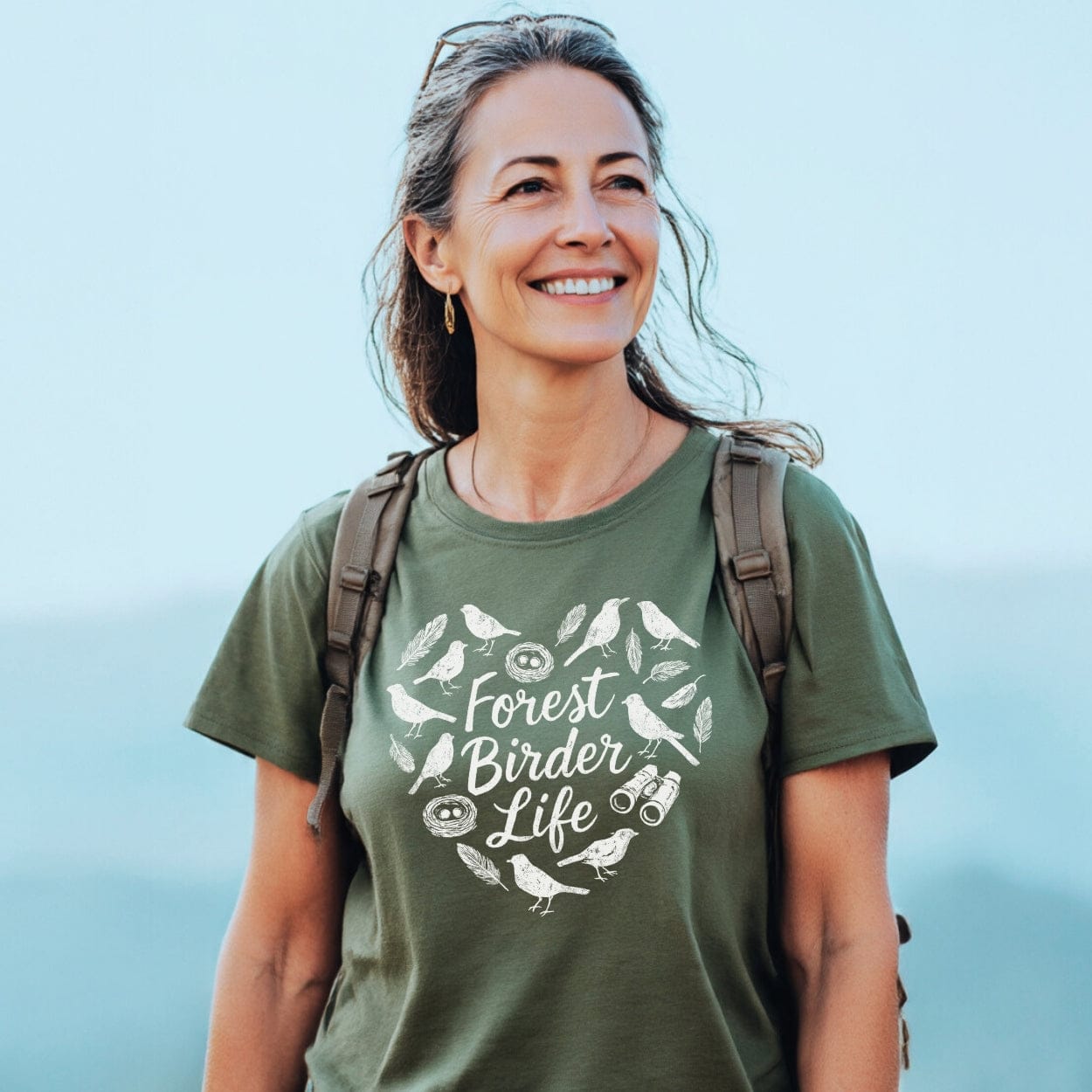 Smiling woman outdoors wearing the Forest Birder Life T-Shirt, featuring a heart design with bird silhouettes and binoculars, perfect for nature hikes. Carries a backpack, embodying outdoor adventure spirit.