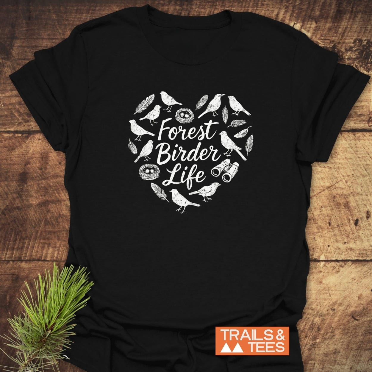 Forest Birder Life T-Shirt with white hand-drawn design of birds, feathers, nests, and binoculars in a heart shape. Features TRAILS & TEES logo and pine sprig, embodying outdoor adventure.