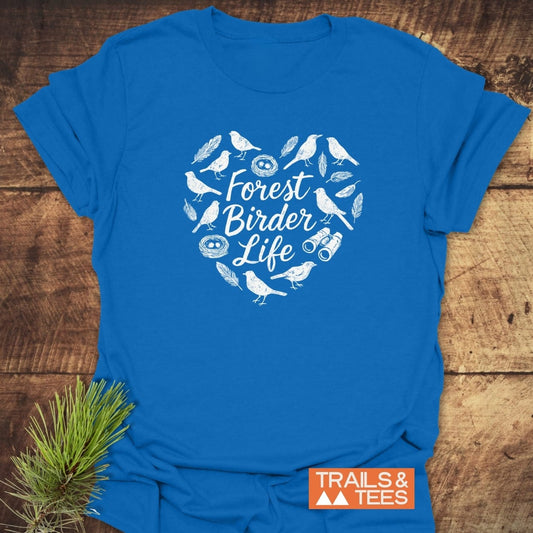 Forest Birder Life T-Shirt featuring a heart-shaped design of bird silhouettes, feathers, and binoculars on a blue cotton fabric, placed on wood. Includes TRAILS & TEES logo and pine sprig.