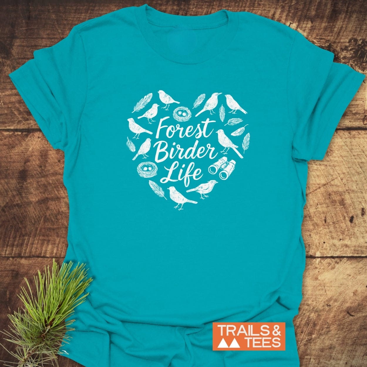 Forest Birder Life T-Shirt on wood, displaying a heart-shaped wreath of bird silhouettes, nests, and binoculars. Features orange TRAILS & TEES label, pine sprig, emphasizing outdoor adventure spirit.
