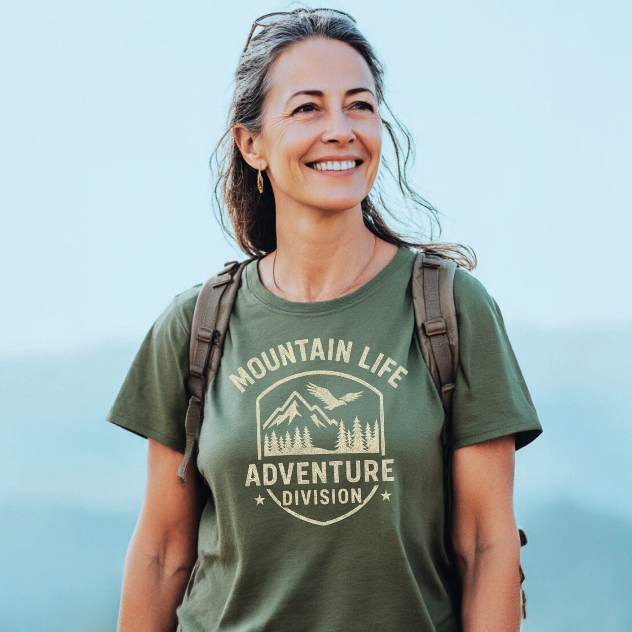 Middle-aged woman outdoors in a Mountain Life T-Shirt, showcasing its comfort and adventure-ready design with a logo. She wears a backpack, embodying the spirit of outdoor exploration.
