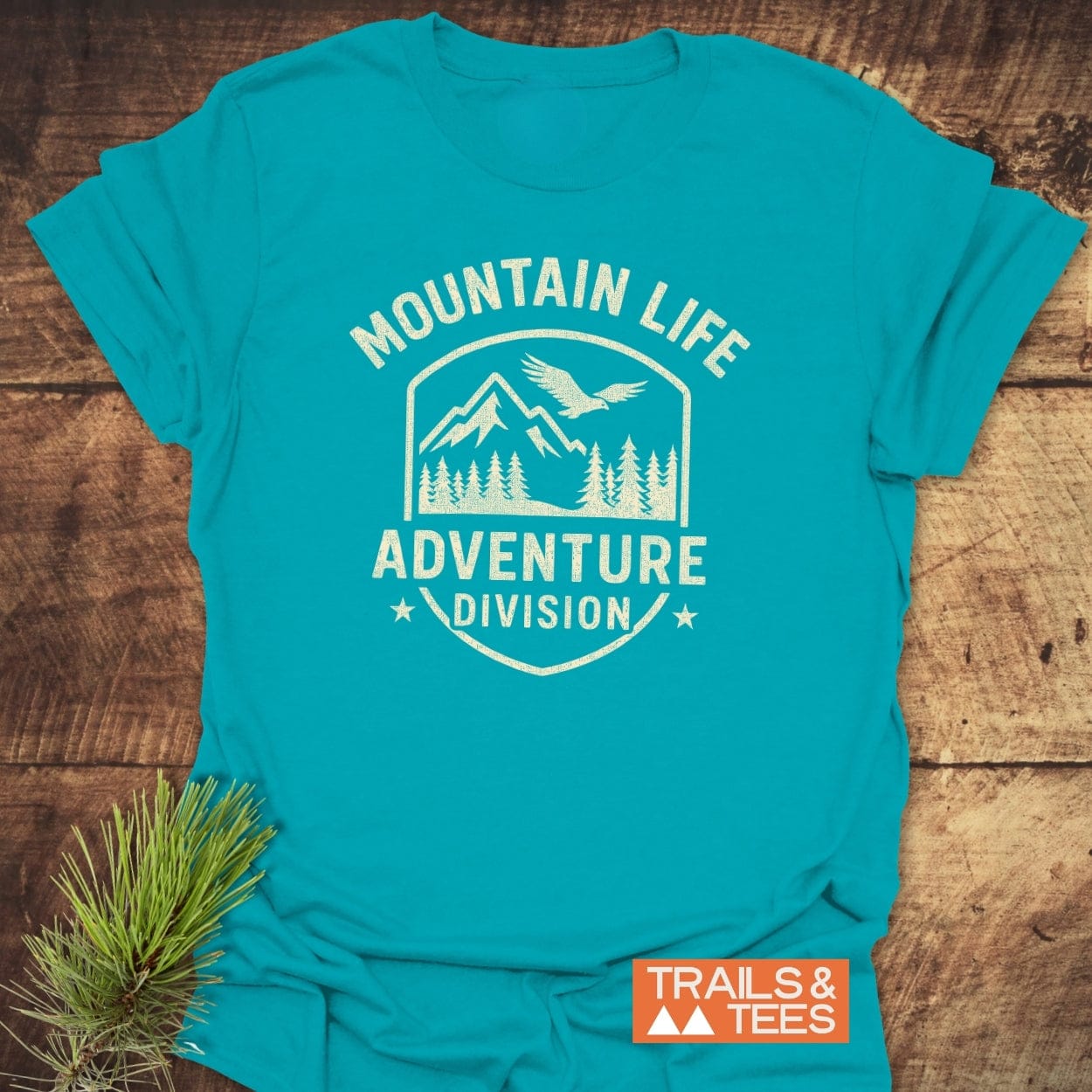 Mountain Life T-Shirt features a distressed white shield graphic with mountains, pine trees, and a bird. Positioned on a rustic wooden surface with a pine sprig, perfect for outdoor enthusiasts.