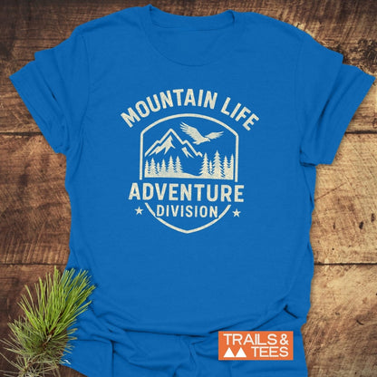 Mountain Life T-Shirt with a distressed emblem featuring mountain peaks, conifer trees, and a bird. Laid on a wooden surface beside a pine sprig, highlighting its adventure-inspired design.