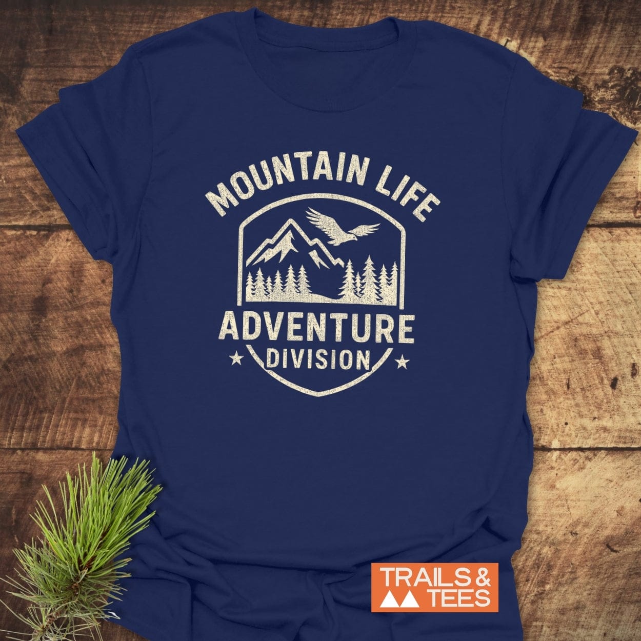 Mountain Life T-Shirt on a wooden surface, featuring a white shield logo with mountains, pine trees, and bird. Includes ADVENTURE DIVISION text. Pine sprig nearby; TRAILS & TEES label visible.