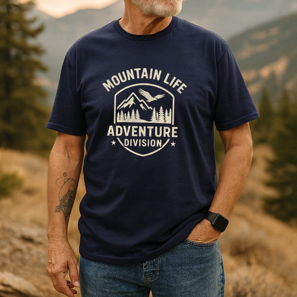 Mountain Life T-Shirt featuring a shield graphic with a mountain peak, eagle, and pine trees. Text reads MOUNTAIN LIFE and ADVENTURE DIVISION. Model wears it with jeans, showcasing outdoor spirit.