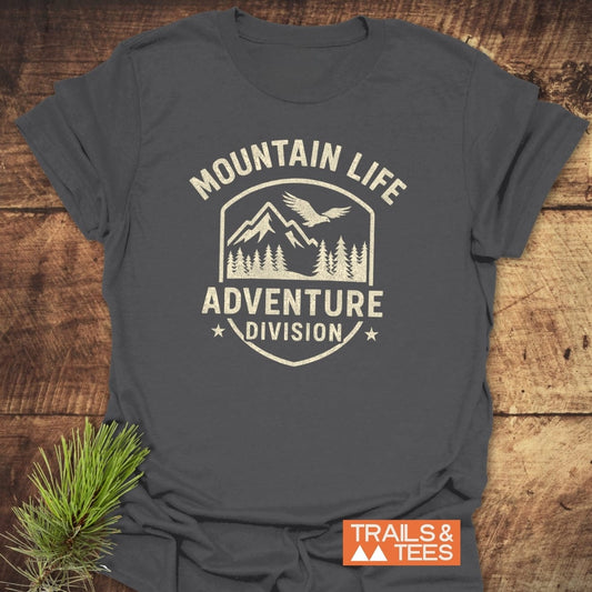 Mountain Life T-Shirt featuring a cream graphic of mountain peaks, trees, and eagle. Text “MOUNTAIN LIFE ADVENTURE DIVISION” with stars. Labeled “TRAILS & TEES,” perfect for outdoor enthusiasts.
