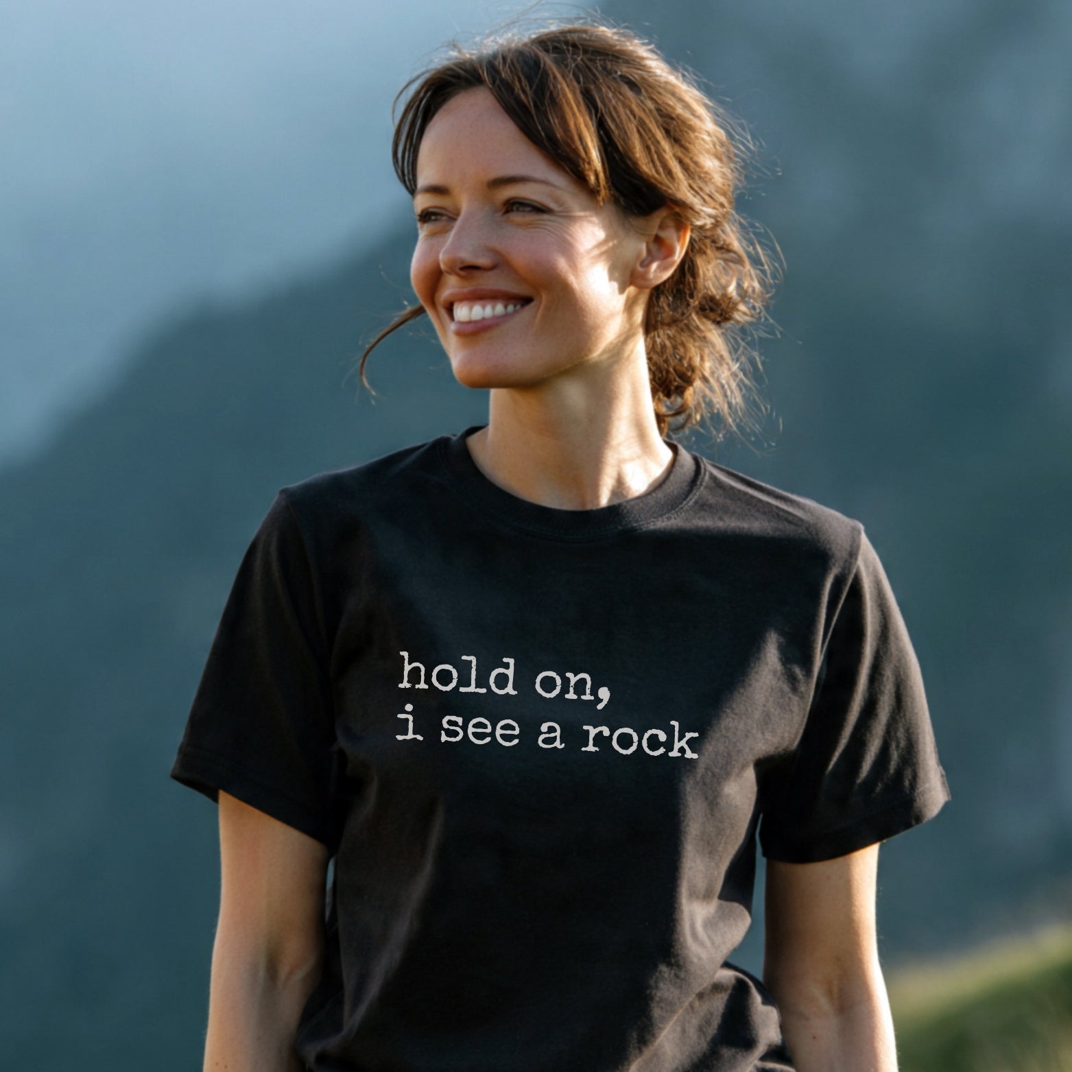 Woman wearing Hold On I See A Rock T-Shirt with a black background and white typewriter-style text, standing outdoors with a blurred nature backdrop, smiling, showcasing its casual, adventure-ready design.