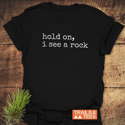 Hold On I See A Rock T-Shirt laid flat, featuring a white typewriter-style phrase. Accompanied by a pine sprig and the Trails & Tees logo, highlighting its outdoor-themed design.