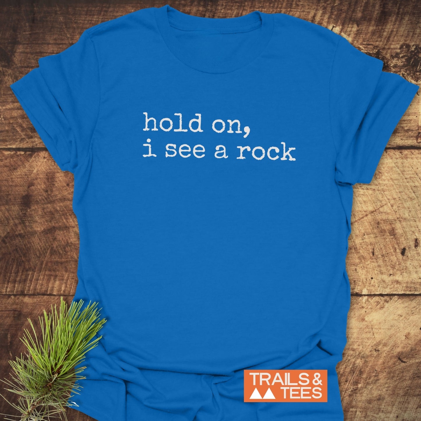 Hold On I See A Rock T-Shirt on wooden surface, features white typewriter-style text; nearby pine sprig. Includes orange “TRAILS & TEES” logo. Made from soft, durable ring-spun cotton.