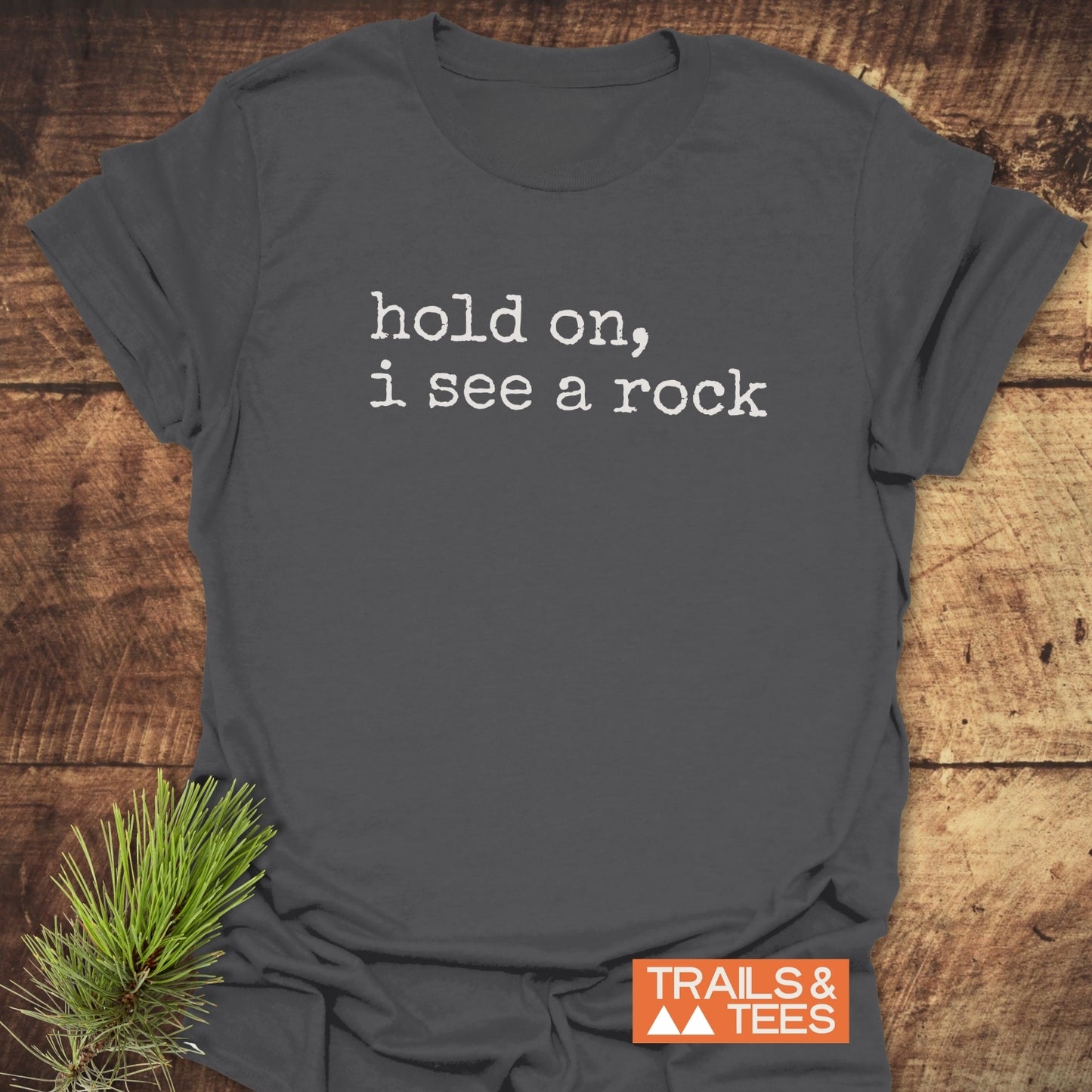Hold On I See A Rock T-Shirt features white typewriter-style text on a dark gray crew-neck, laid flat with a pine sprig. Includes an orange TRAILS & TEES label.
