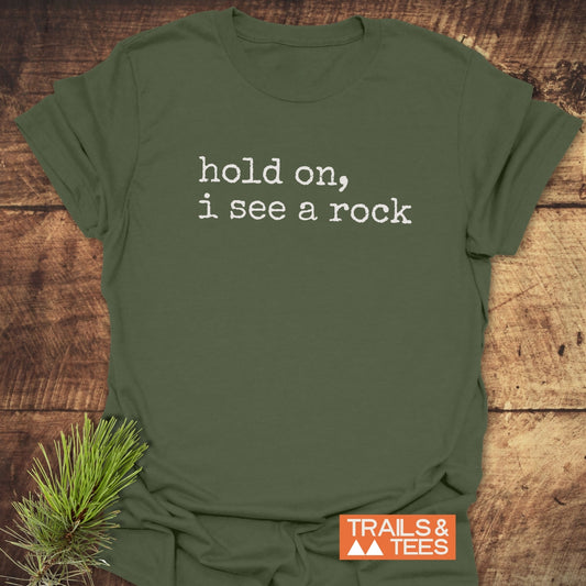Hold On I See A Rock T-Shirt displayed on wood, featuring white typewriter-style text. Includes TRAILS & TEES orange logo with mountain icons. Accented with a pine bough for a nature-inspired touch.