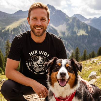 Bearded man wearing Hiking? Australian Shepherd T-Shirt crouches on mountain trail beside a happy dog with a red bandana, set against forested slopes and distant rocky peaks.