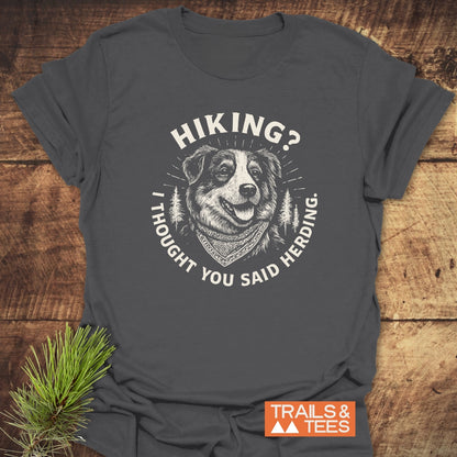 Hiking? Australian Shepherd T-Shirt featuring a cheerful herding dog with bandana and pine trees design. Laid on wood, includes TRAILS & TEES logo, highlighting adventure-ready, comfortable cotton.