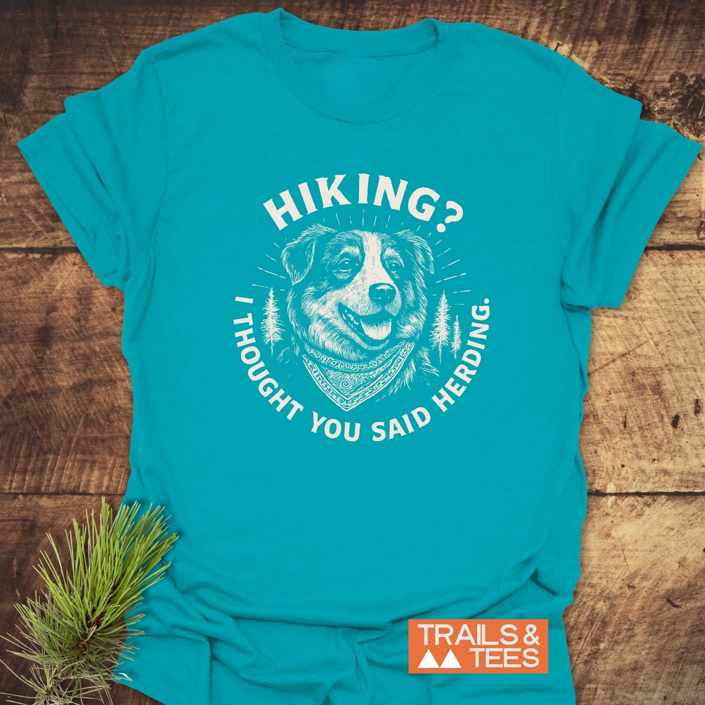 Hiking? Australian Shepherd T-Shirt featuring a happy dog graphic with a pun slogan, laid flat on a wooden surface next to a pine branch.