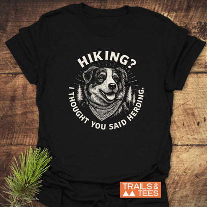 Hiking? Australian Shepherd T-Shirt featuring a happy Border Collie graphic on black fabric, laid on rustic wood with pine sprig. Includes 'TRAILS & TEES' logo, highlighting outdoor adventure spirit.
