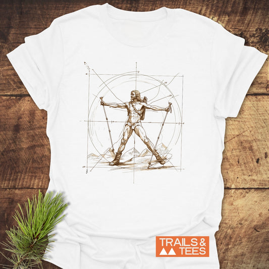 Vitruvian Hiker Da Vinci Style T-Shirt featuring a sepia sketch of a hiker with trekking poles and mountains, set against measuring lines. Trails & Tees logo visible, laid on a wooden surface.