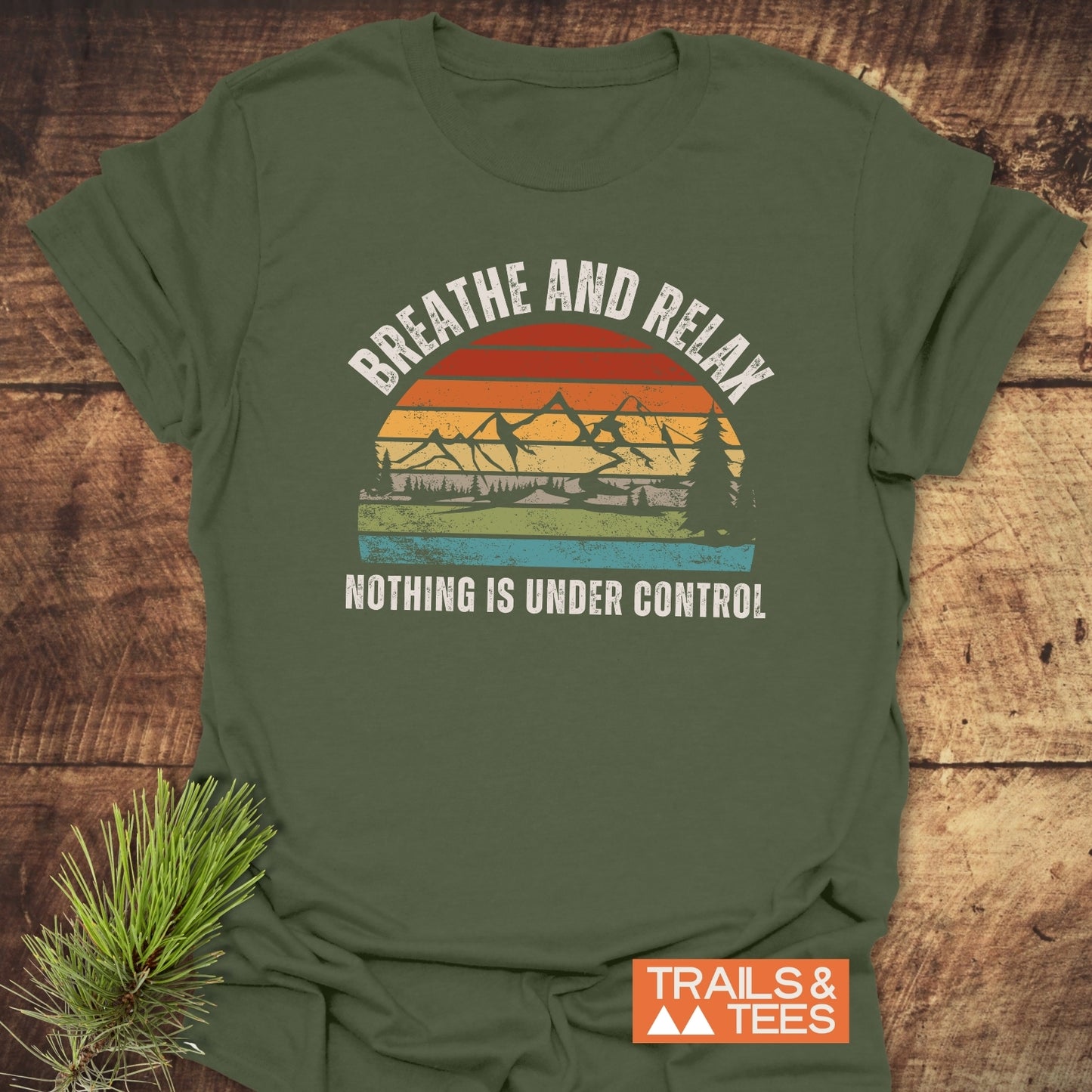 Moss-green Breathe And Relax Funny T-Shirt featuring a vintage sunset graphic with pine trees and mountains, laid on wood with pine branch, promoting outdoor comfort and adventure.