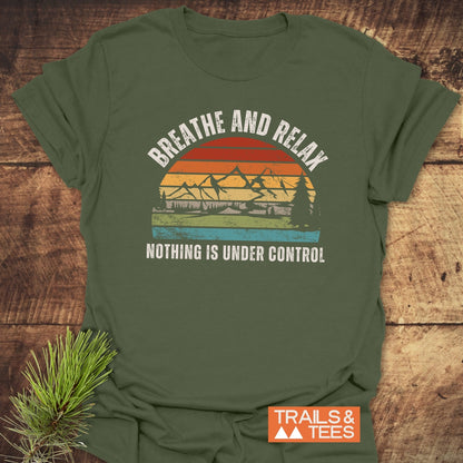 Moss-green Breathe And Relax Funny T-Shirt featuring a vintage sunset graphic with pine trees and mountains, laid on wood with pine branch, promoting outdoor comfort and adventure.