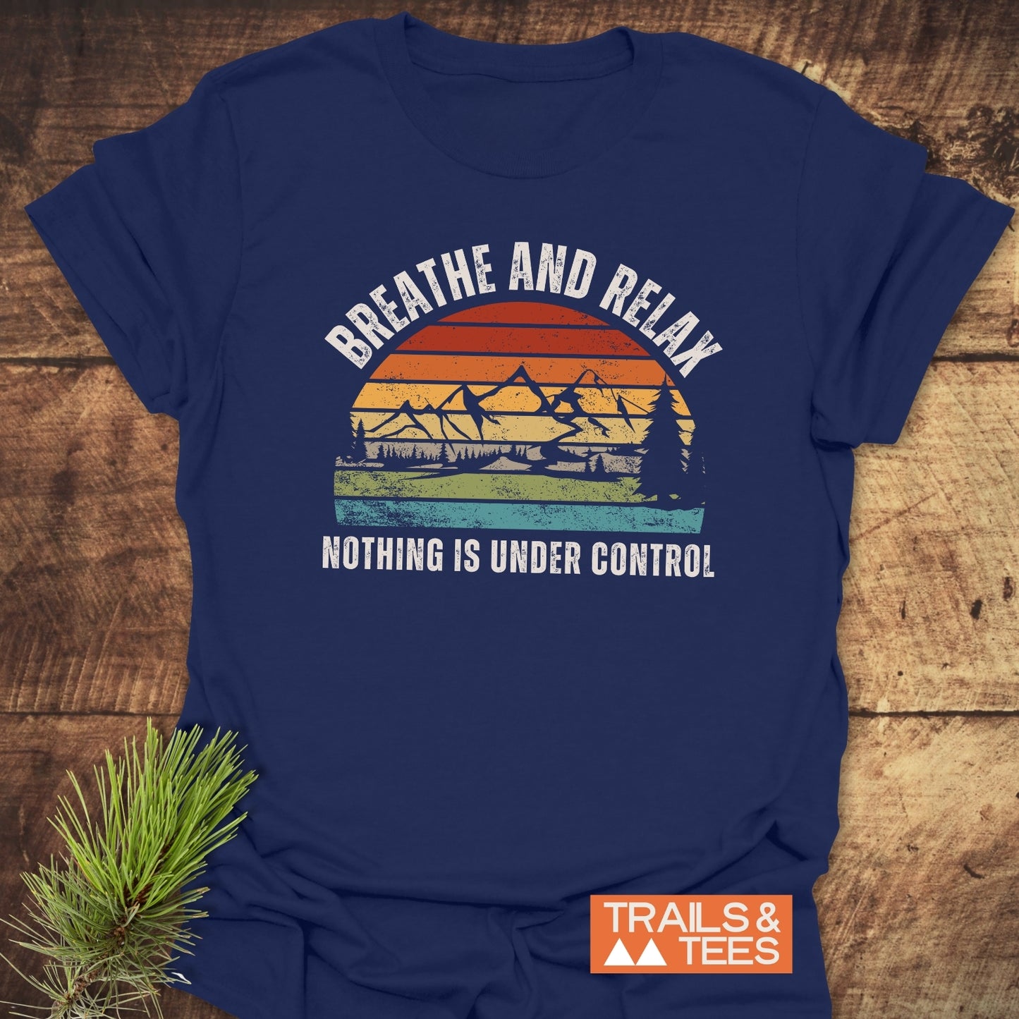 Navy t-shirt with vintage graphic and “BREATHE AND RELAX NOTHING IS UNDER CONTROL” text, featuring Trails & Tees logo, lying on wood with pine sprig.