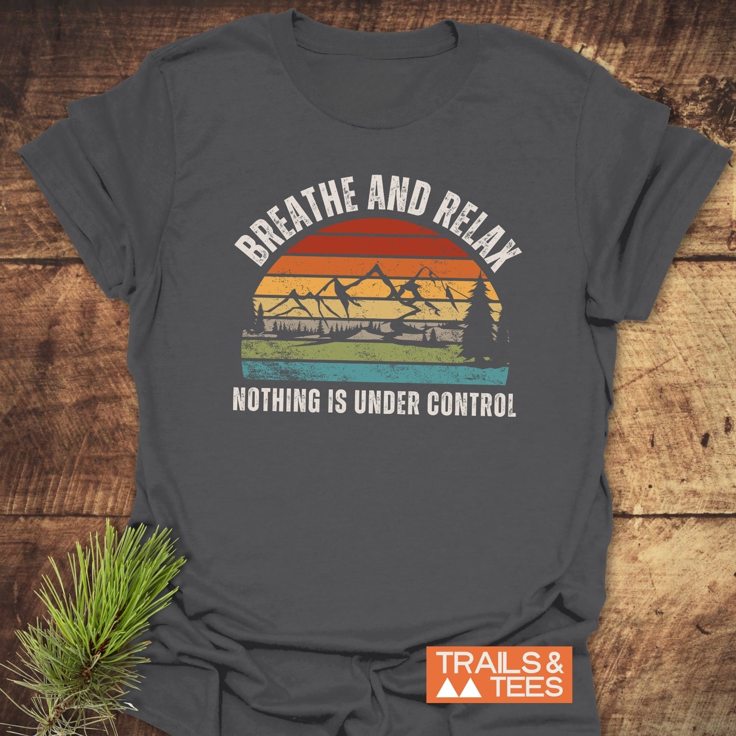Charcoal-gray Breathe And Relax Funny T-Shirt featuring a vintage sunset graphic with mountains, trees, and a lake, emphasizing outdoor adventure. Includes TRAILS & TEES logo; pine sprig accents the display.