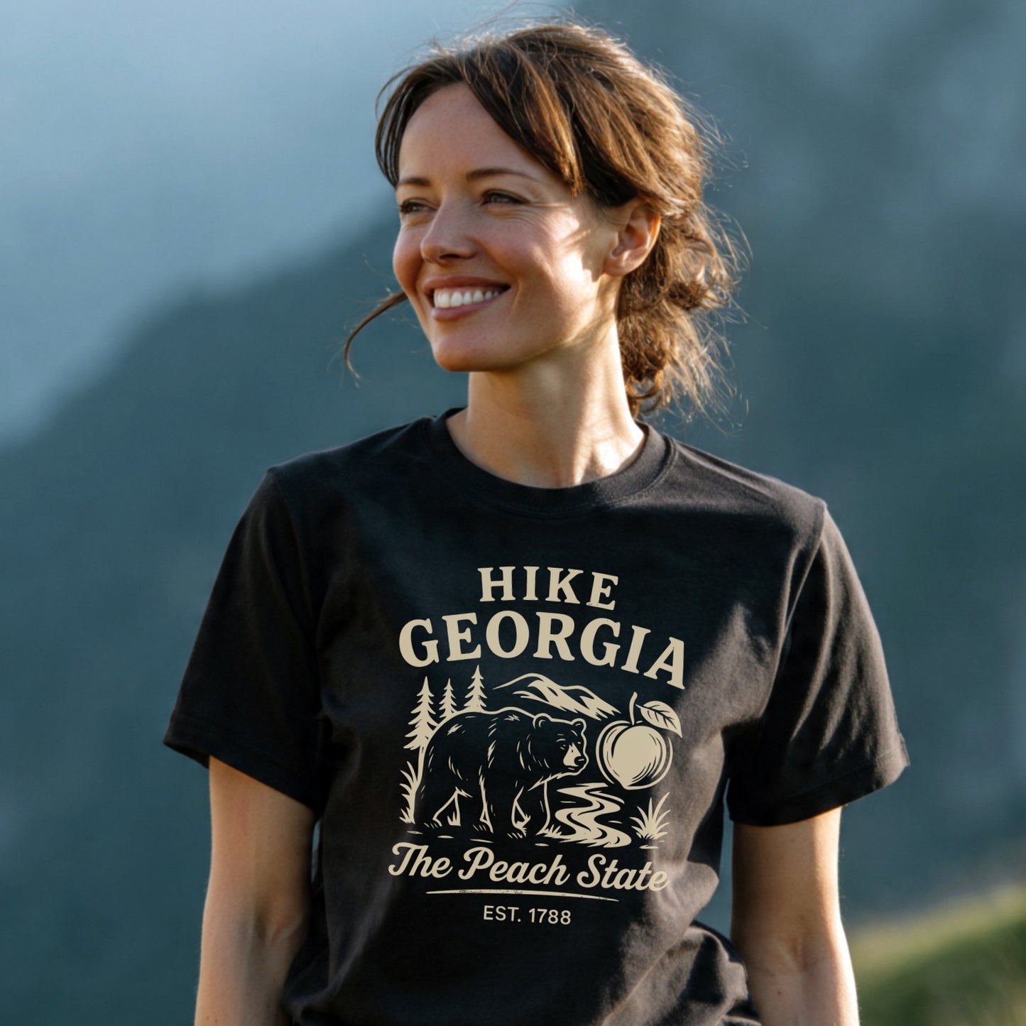 Woman outdoors wearing Hike Georgia T-Shirt featuring a bear, pine trees, stream, mountain, and peach design. The shirt embodies comfort with soft cotton, pre-shrunk fit, and durable prints.