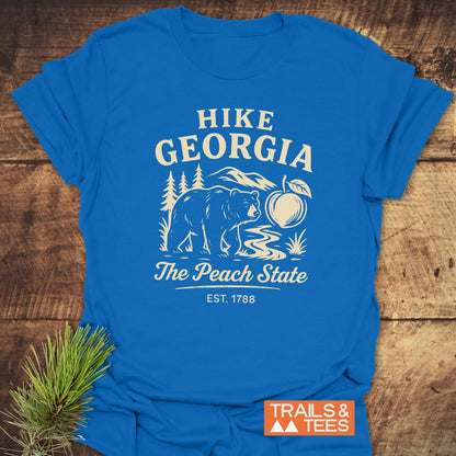 Hike Georgia T-Shirt featuring a bear, stream, and mountains design with The Peach State and EST. 1788 text, laid on wood, showcasing outdoor spirit and durable, soft cotton craftsmanship.