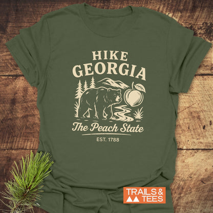 Hike Georgia T-Shirt featuring a bear, mountain stream, pine trees, and a peach graphic, with The Peach State and EST. 1788 text. Includes TRAILS & TEES logo and pine needle sprig.