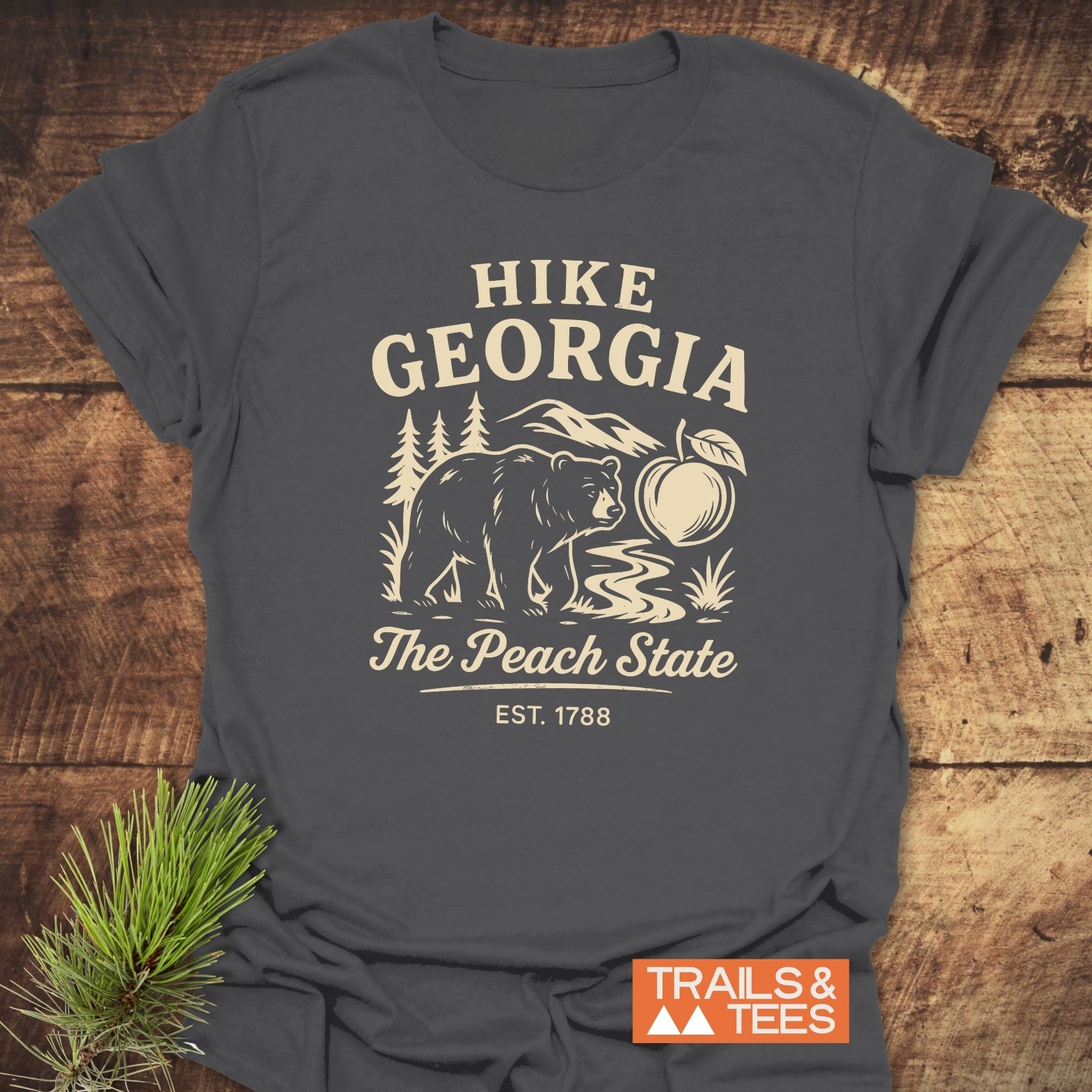 Hike Georgia T-Shirt featuring a vintage bear graphic with mountains, a peach, and The Peach State. Laid on wood with pine sprig, highlighting its trail-ready, soft cotton design by Trails & Tees.