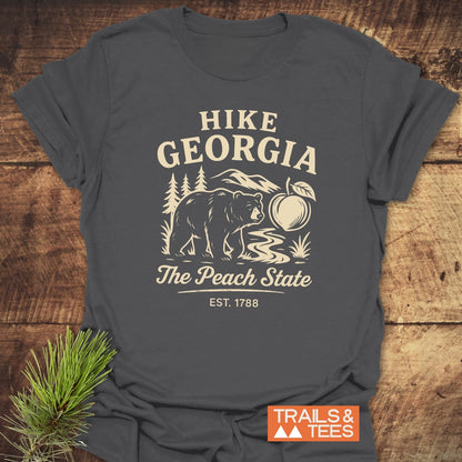Hike Georgia T-Shirt featuring a vintage bear graphic with mountains, a peach, and The Peach State. Laid on wood with pine sprig, highlighting its trail-ready, soft cotton design by Trails & Tees.