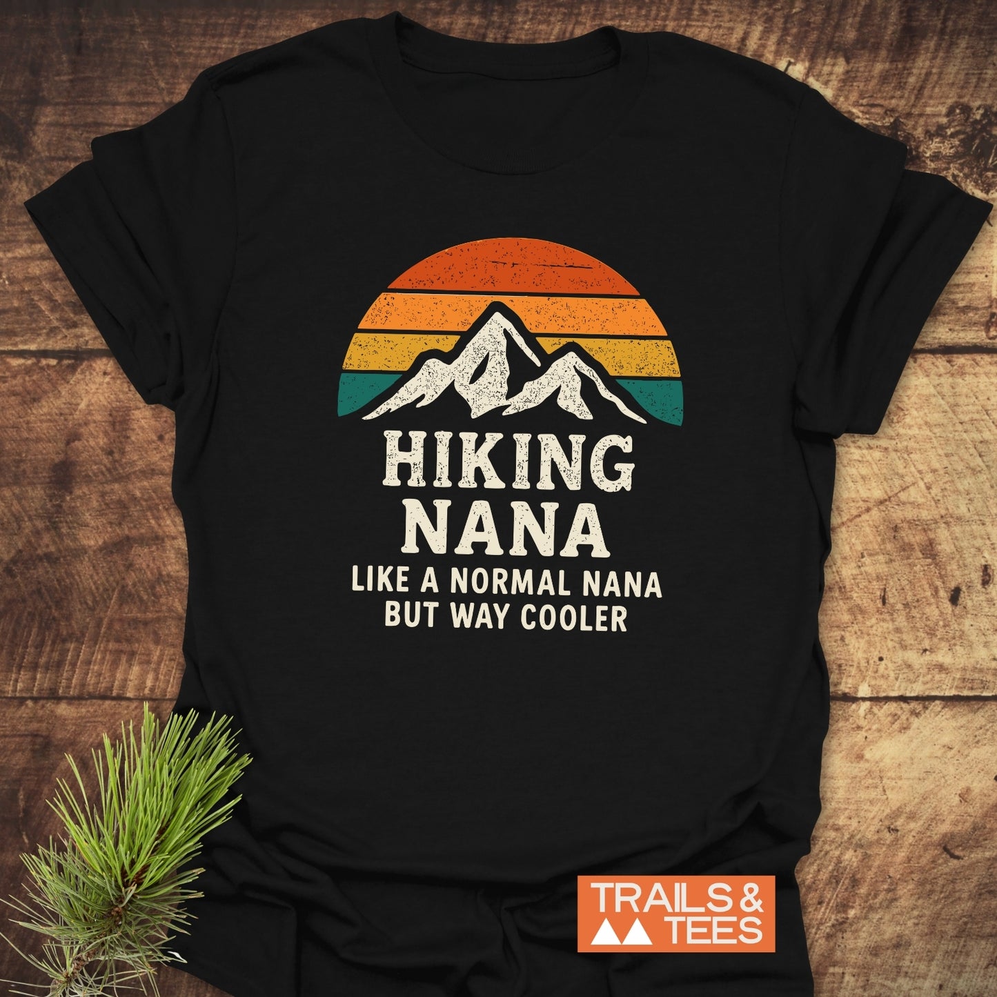 Black Hiking Nana Funny T-Shirt featuring a retro sunset and mountain graphic with bold text, laid flat on wood, highlighting comfort and design. Pine sprig accentuates the outdoor theme.