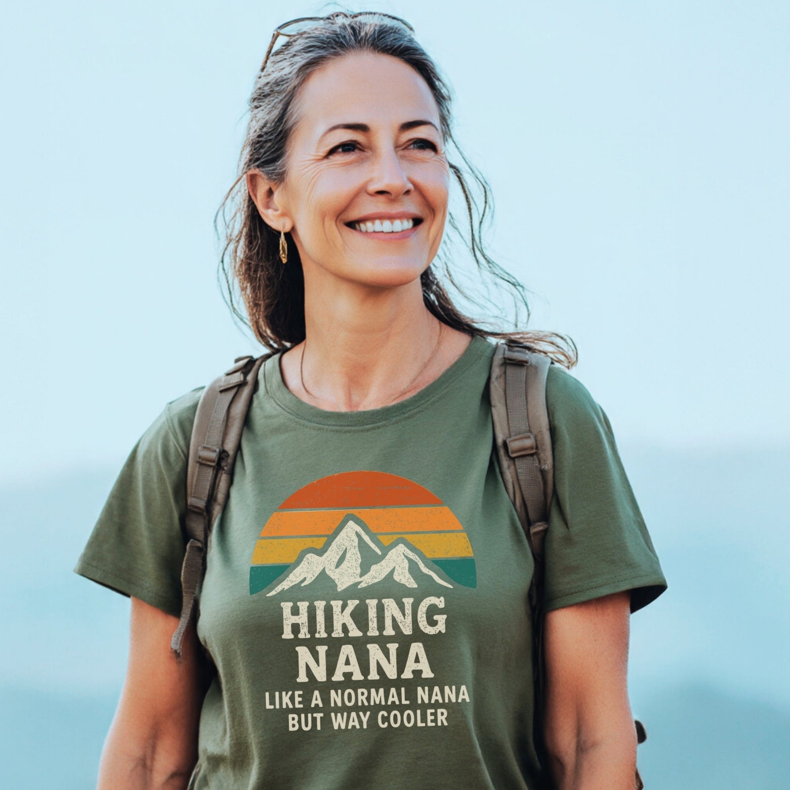 Middle-aged woman outdoors in an olive-green Hiking Nana Funny T-Shirt featuring a mountain silhouette against a sunset, expressing a love for hiking and adventure.